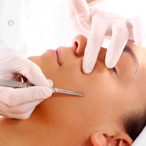 A dermaplaning blade being drawn across a reclined client’s cheek