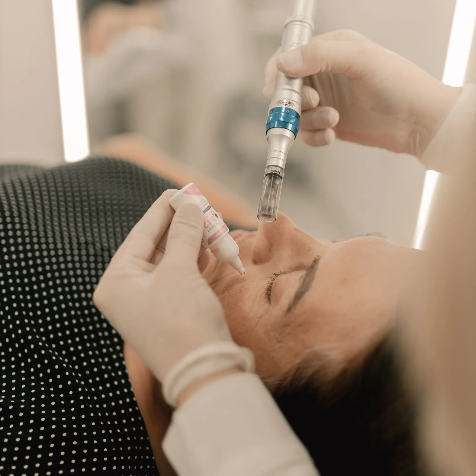 A microneedling pen held over a patient’s cheek while serum is applied, in the practice’s slate duotone