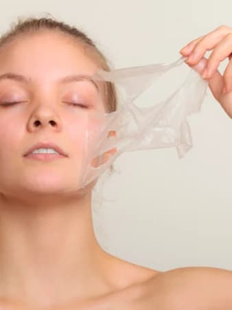 A woman with her eyes closed lifting a translucent treatment mask away from her cheek