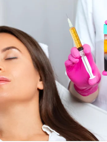 A gloved hand holding a syringe of separated golden plasma beside a woman reclined for treatment